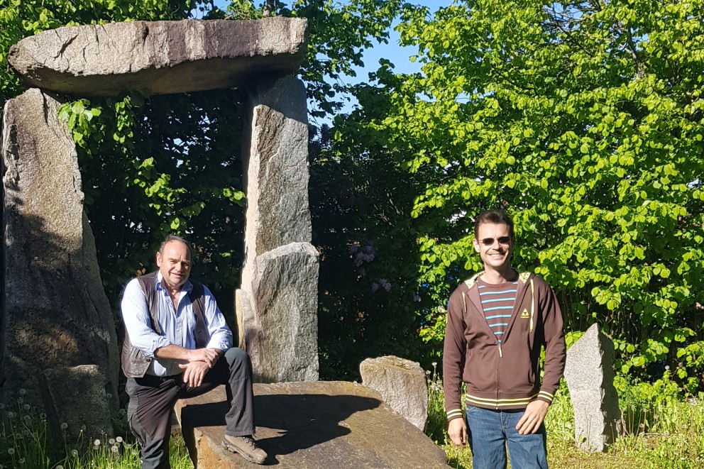 Zwei Männer vor einem Torbogen aus Stein in einem Garten.