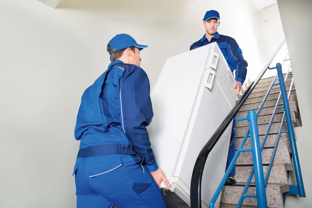 Zwei Umzugshelfer tragen einen Kühlschrank die Treppe hoch.