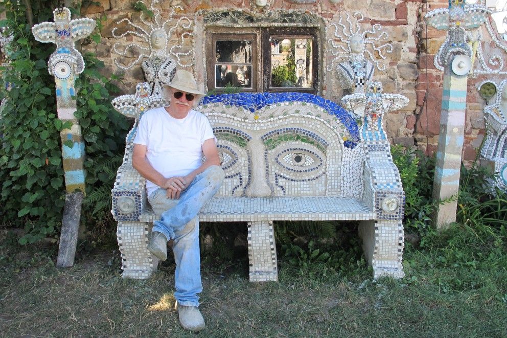 
				Otfried Culmann ruht aus auf der vollendeten «Kopf Bank», links und rechts der Bank Stelen mit Eulen, ebenfalls aus Mosaik gefertigt

			