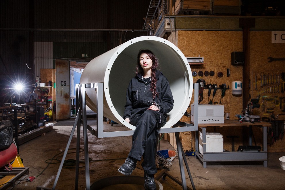 Une femme est assise dans un grand tube sur un cadre métallique dans un atelier.