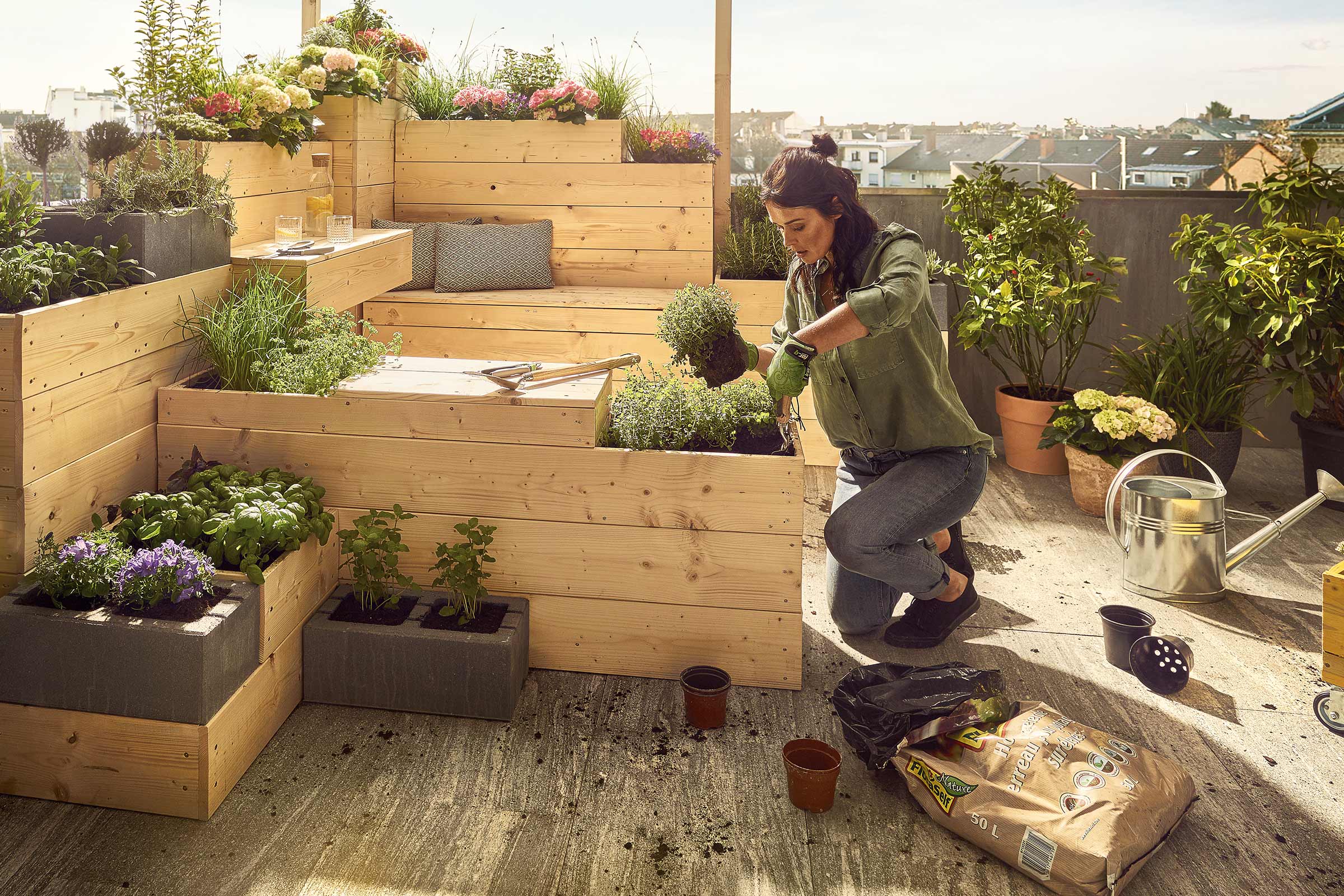 Frau bepflanzt Hochbeet auf einer Dachterrasse mit Gartenerde, Kräutern und Blumen.