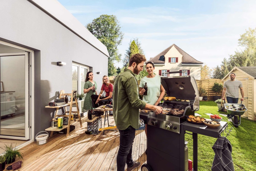 Personen grillieren im Garten auf einem Grill mit Seitenablage und geniessen Getränke.
