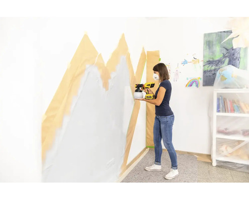 Frau sprüht Wandfarbe mit Farbsprühsystem auf eine Wand, die mit Malerkrepp abgeklebt ist.