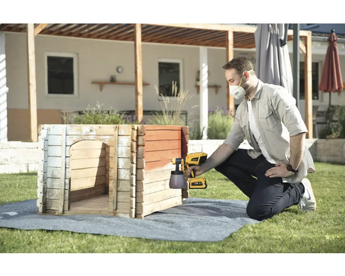 Mann streicht mit einer Farbspritzpistole ein Hundehaus im Garten.