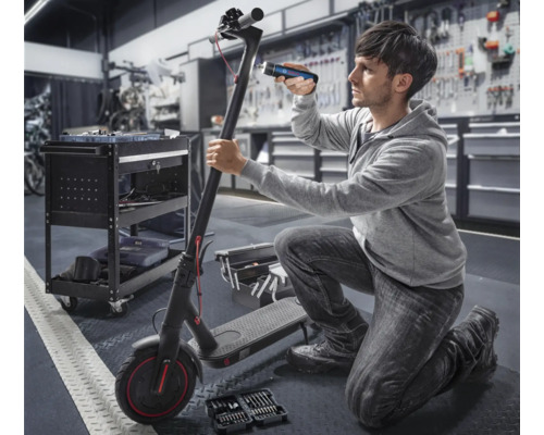 Un homme répare une trottinette électrique avec des outils et une lampe dans un atelier.