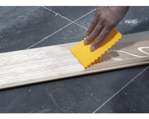 Eine Person verteilt Klebstoff mit einem Spachtel auf einem Holzbrett.