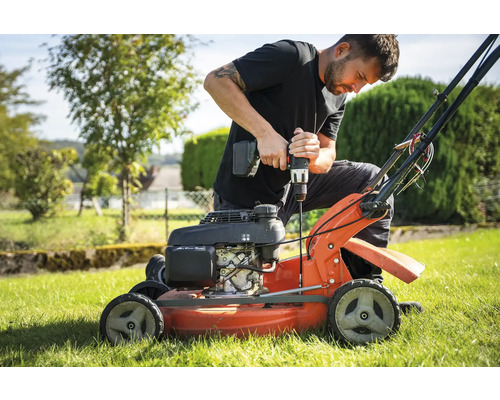 Un homme répare une tondeuse à gazon avec une visseuse sans fil dans le jardin.