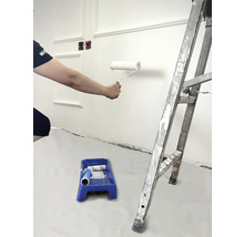 Person streicht Wand mit Farbroller, Farbschale und Leiter im Raum.