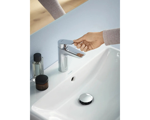 Aufnahme eines Badezimmers mit einem weißen Waschbecken, einem Chrom-Wasserhahn, einer Hand, die den Wasserhahn betätigt, und zwei Behältern