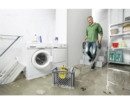 Cave inondée avec machine à laver, sèche-linge et pompe submersible dans un panier. Un homme en bottes en caoutchouc traverse l''eau.