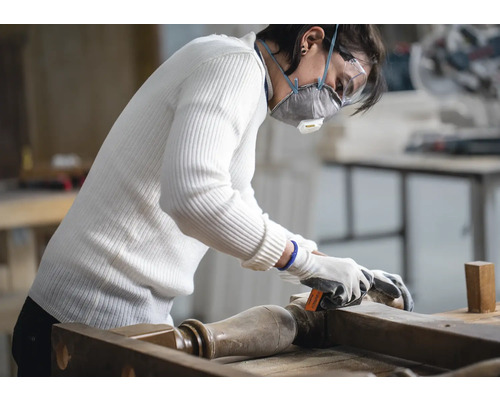 Une personne ponce un meuble avec du papier de verre, portant un masque de protection et des lunettes de sécurité.