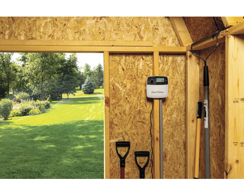 Scène d''un système d''irrigation de jardin avec un contrôleur Rain Bird et des outils de jardin dans une cabane en bois, vue sur une pelouse avec irrigation active
