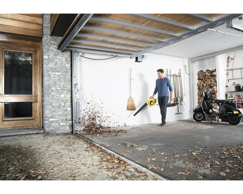 Un homme utilise un souffleur de feuilles dans un garage pour enlever les feuilles. À l''arrière-plan, on aperçoit un scooter et des piles de bois.