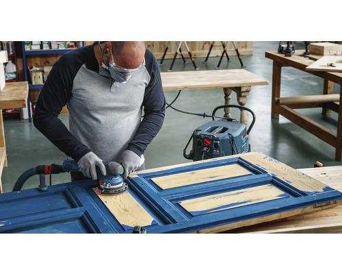Eine Person schleift eine blaue Holztür mit einem Exzenterschleifer, der an einen Staubsauger angeschlossen ist.
