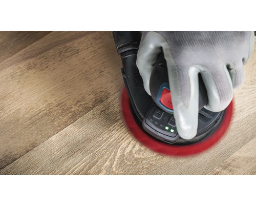 Une main gantée utilise une ponceuse excentrique sur un plancher en bois.