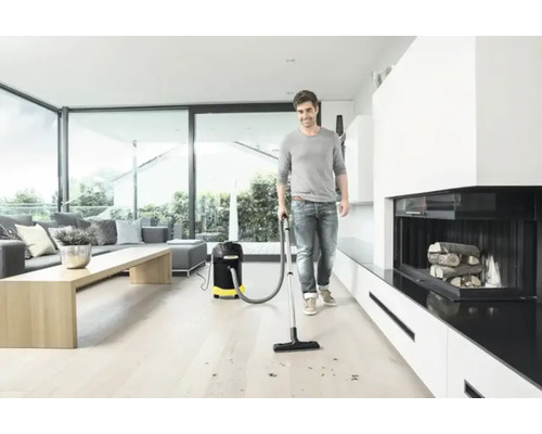 Un homme aspire des copeaux de bois avec un aspirateur multifonction dans un salon lumineux.