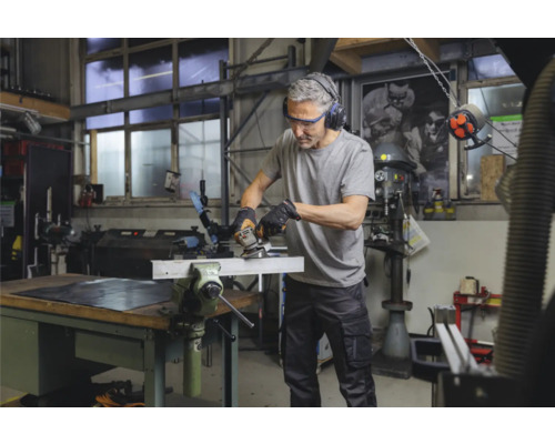 Un homme travaille dans un atelier avec une meuleuse d''angle sur une pièce métallique serrée dans un étau. Il porte des lunettes de protection et un casque antibruit.