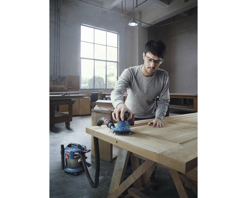 Un homme ponce une porte en bois avec une ponceuse excentrique et un aspirateur dans un atelier.