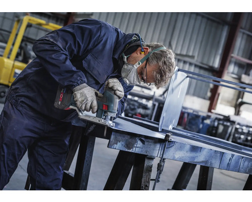 Homme avec masque de protection, gants et protections auditives sciant une pièce métallique avec une scie sauteuse sans fil