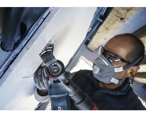 Handwerker bei der Arbeit mit einem Multifunktionswerkzeug an einer Decke, ausgestattet mit Schutzbrille und Staubschutzmaske