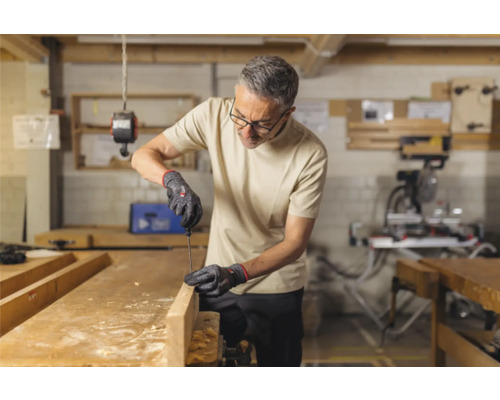 Un homme travaille avec un tournevis et des gants sur une planche de bois dans un atelier.
