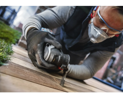 Ein Mann mit Schutzbrille, Atemschutz und Handschuhen arbeitet mit einem Multifunktionswerkzeug an einem Holzbrett.