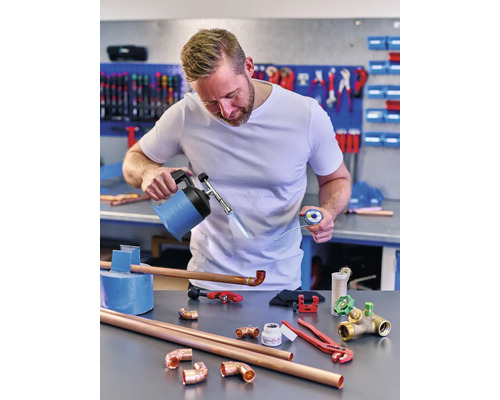 Un homme soude des tubes de cuivre avec un brûleur, de la soudure et divers outils sur un établi.