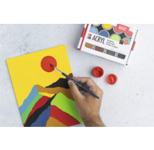 Une personne peint un motif de montagne abstrait avec des peintures acryliques sur une toile, à côté d''une boîte de peintures acryliques.