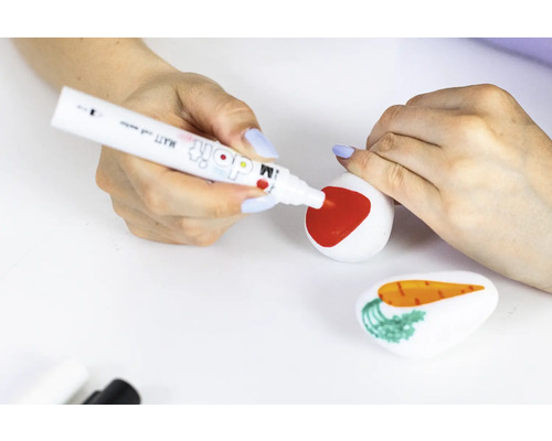 Eine Person bemalt einen Stein mit einem roten Stift, während ein weiterer Stein mit einer Karotte im Vordergrund liegt.