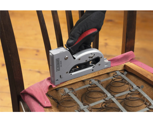 Eine Hand mit Handschuh befestigt Stoff mit einem Handtacker auf einem Stuhl.