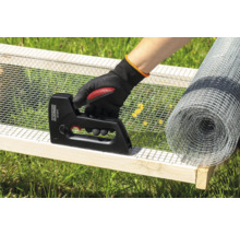 Eine Person befestigt Drahtgeflecht mit einem Tacker an einem Holzrahmen, mit einer Rolle Drahtgeflecht im Hintergrund.