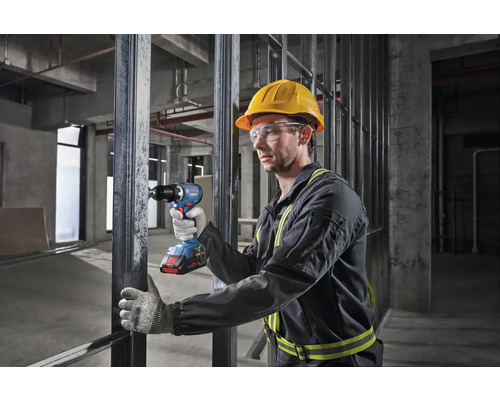 Ein Arbeiter mit Schutzhelm und Schutzbrille benutzt einen Akku-Bohrschrauber auf einer Baustelle.