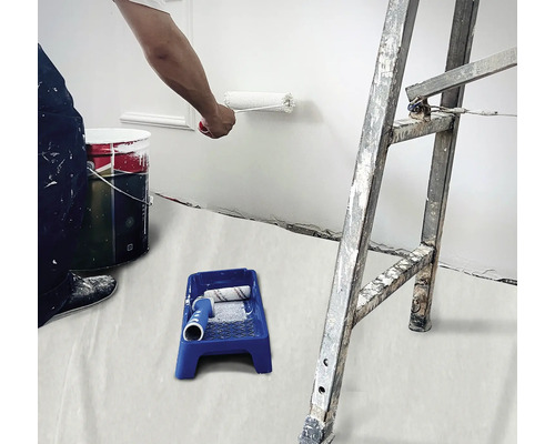 Un homme peint un mur avec un rouleau à peinture, à côté de lui un pot de peinture, un bac à peinture et une échelle.