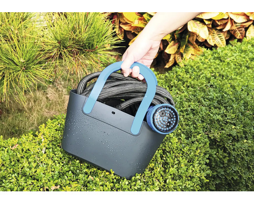 Une personne tient un tuyau d''arrosage avec pistolet d''arrosage dans un panier devant des buissons.