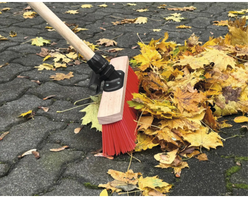 Eine Kehrmaschine mit Holzstiel und roten Borsten kehrt Laub auf einem Pflasterweg.
