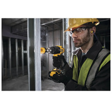 Un homme portant un casque de protection, des lunettes de sécurité et une protection auditive perce un trou dans un mur à l''aide d''une perceuse-visseuse sans fil jaune.