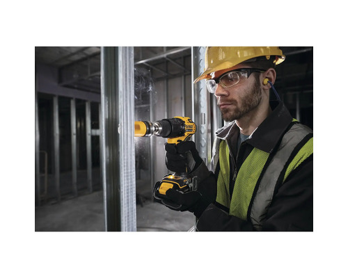 Ein Mann mit Schutzhelm, Schutzbrille und Gehörschutz bohrt mit einem gelben Akku-Bohrschrauber ein Loch in eine Wand.