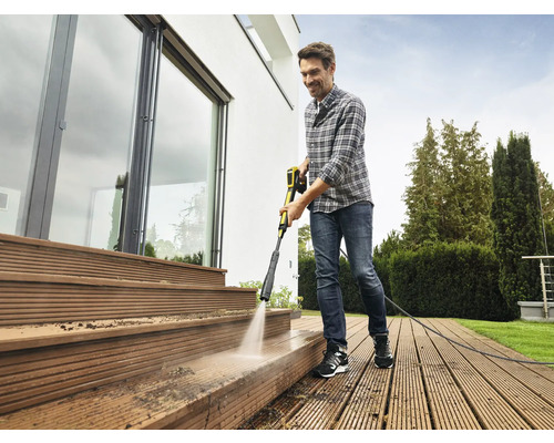 Un homme nettoie une terrasse en bois devant une maison avec un nettoyeur haute pression.