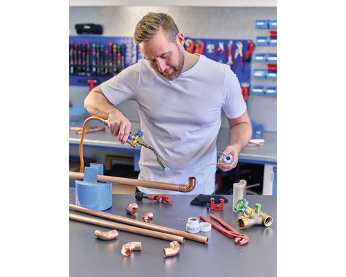 Un installateur soude des tuyaux en cuivre avec un chalumeau, tandis que divers outils et pièces de tuyaux sont posés sur l''établi.