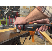 Eine Person benutzt ein Multifunktionswerkzeug der Marke Worx, um ein Stück Holz zu bearbeiten. Das Werkzeug ist schwarz und orange.