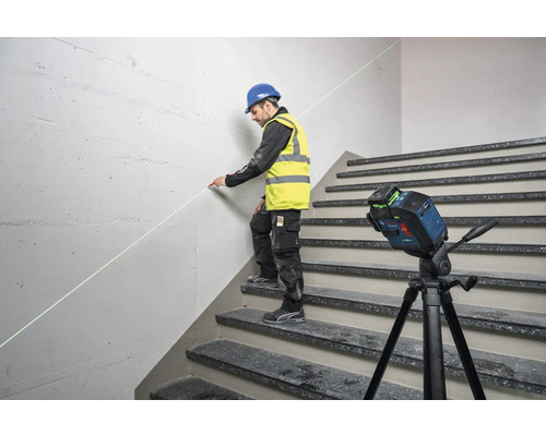 Ein Mann mit Schutzhelm und Warnweste benutzt einen Linienlaser auf einer Treppe, um eine Markierung an einer Wand anzubringen.