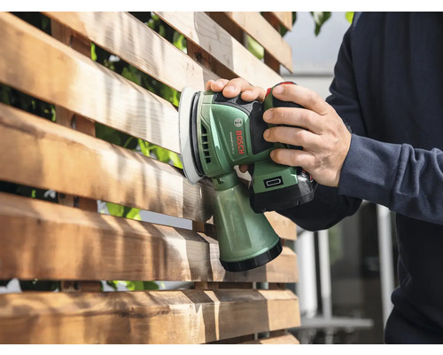 Eine Person schleift mit einem Akku-Exzenterschleifer einen Holzzaun.