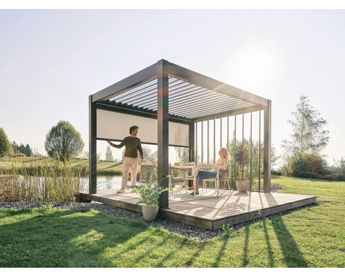 Pergola de terrasse autoportante avec toit à lames, store latéral et deux personnes dans le jardin.