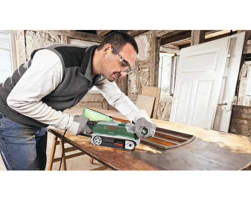 Un homme ponce une porte en bois avec une ponceuse à bande. Il porte des lunettes de protection et un casque antibruit.