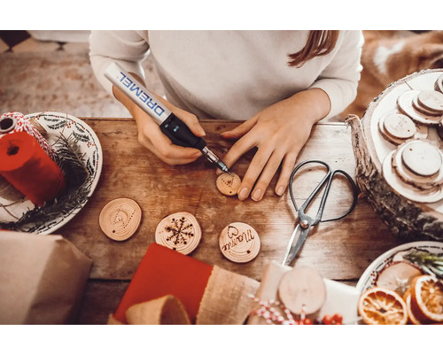 Une femme grave des étiquettes cadeaux en bois avec un outil Dremel sur une table entourée de décorations de Noël.