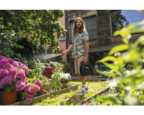 Mann bewässert Garten mit Schlauch und Schlauchwagen vor Haus