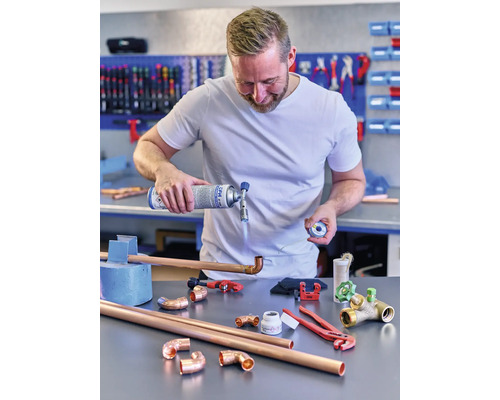 Un homme soude des tuyaux en cuivre avec un brûleur à gaz et des outils sur une table.