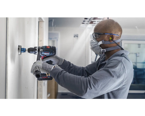 Un artisan perce un trou dans un mur avec une scie cloche. Il porte un masque de protection et des lunettes de sécurité. Une perceuse du fabricant Bosch est en service.