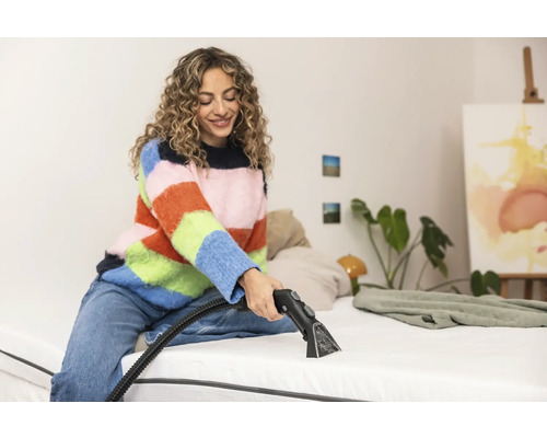 Une femme nettoie un matelas avec un aspirateur injecteur-extracteur dans une chambre lumineuse.