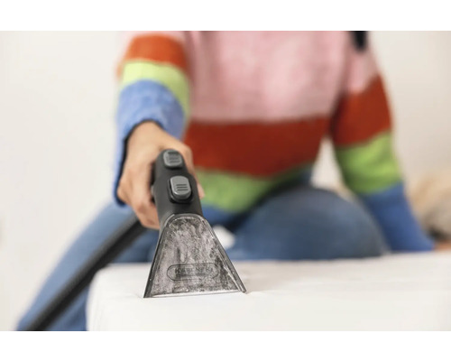 Gros plan d''une personne utilisant un nettoyeur de tissus d''ameublement sur un matelas. L''appareil est doté d''un embout transparent et de deux boutons gris.